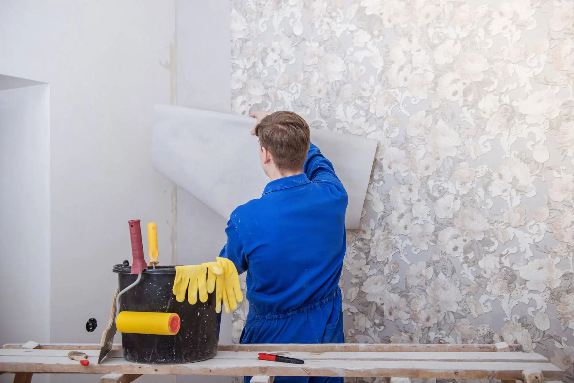 Worker in blue coveralls lifting a loose sheet from a pale floral-patterned wall beside a bucket of tools.